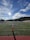 A sports field with green grass and a goalpost is in the foreground. In the distance, a group of people is gathered by the sideline. There are mountains in the background under a blue sky with scattered clouds. The sun is shining brightly, casting long shadows across the field.