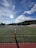 A sports field with green grass and a goalpost is in the foreground. In the distance, a group of people is gathered by the sideline. There are mountains in the background under a blue sky with scattered clouds. The sun is shining brightly, casting long shadows across the field.