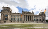 A grand historic building with classical architecture, featuring a large central dome and multiple German flags flying around it. The structure has a series of large columns at its entrance, flanked by symmetrical wings with multiple windows. The foreground includes a patterned stone path and grass lawn leading up to the building.