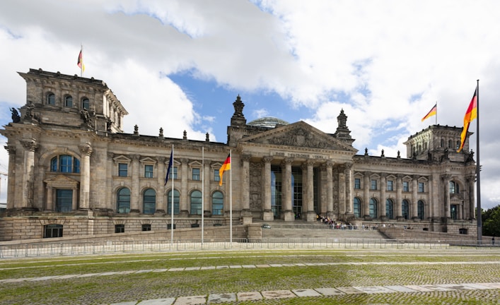A grand historic building with classical architecture, featuring a large central dome and multiple German flags flying around it. The structure has a series of large columns at its entrance, flanked by symmetrical wings with multiple windows. The foreground includes a patterned stone path and grass lawn leading up to the building.