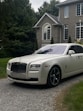 A luxury white Rolls-Royce car is parked on a gravel driveway in front of a large, elegant house with stone walls and large windows. Tall trees and bushes surround the property, creating a lush and private setting.