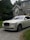 A luxury white Rolls-Royce car is parked on a gravel driveway in front of a large, elegant house with stone walls and large windows. Tall trees and bushes surround the property, creating a lush and private setting.