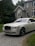 A luxury white Rolls-Royce car is parked on a gravel driveway in front of a large, elegant house with stone walls and large windows. Tall trees and bushes surround the property, creating a lush and private setting.