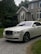 A luxury white Rolls-Royce car is parked on a gravel driveway in front of a large, elegant house with stone walls and large windows. Tall trees and bushes surround the property, creating a lush and private setting.