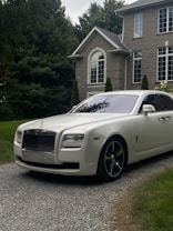 A luxury white Rolls-Royce car is parked on a gravel driveway in front of a large, elegant house with stone walls and large windows. Tall trees and bushes surround the property, creating a lush and private setting.