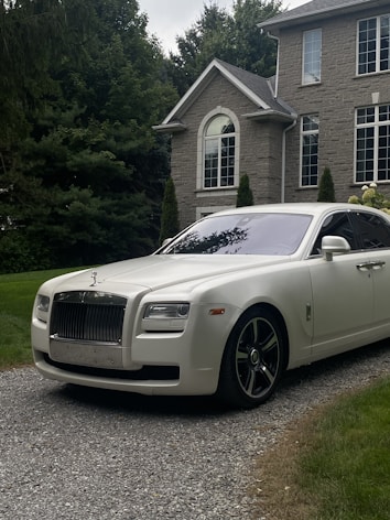 A luxury white Rolls-Royce car is parked on a gravel driveway in front of a large, elegant house with stone walls and large windows. Tall trees and bushes surround the property, creating a lush and private setting.