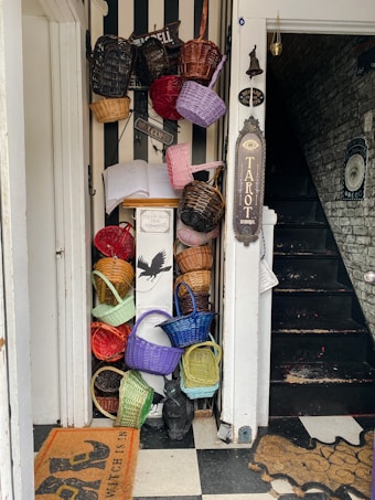 A narrow entryway adorned with an eclectic collection of colorful baskets hanging and stacked against a striped wall. Multiple signs and a tarot sign add a mystical vibe, while a black stairway blends into the background. A raven statue sits beside the stacked baskets, and woven doormats are placed on a checkered floor.
