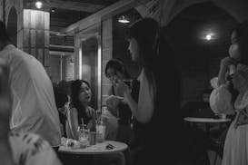 A dimly lit café with several young people gathered around small tables. Some are engaged in conversation, while one person uses a laptop in the background. There are visible drinks on the tables, and a few patrons are dressed in semi-formal attire.