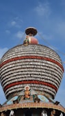 A large, ornately decorated temple chariot featuring a dome with multiple layers of intricate patterns and figures. The structure includes a statue of a deity and is adorned with colorful details, including red and white bands. The background is a clear blue sky.