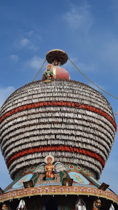 A large, ornately decorated temple chariot featuring a dome with multiple layers of intricate patterns and figures. The structure includes a statue of a deity and is adorned with colorful details, including red and white bands. The background is a clear blue sky.