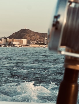 A coastal scene featuring a vast expanse of ocean water with waves, a blurred foreground element of a metallic structure resembling a boat railing or equipment. In the background, there is a hilly landscape with a small structure at the top, likely an antenna or tower, and several modern buildings along the shore.