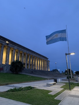 A large neoclassical building with columns is illuminated at dusk. A flagpole with an Argentine flag flutters in the breeze, set against a deep blue sky. The foreground features a pathway with grass and benches, and there are streetlights along the path.