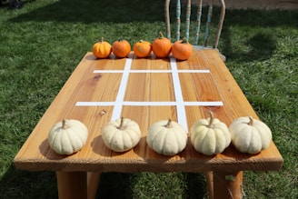 A wooden table outdoors with a tic-tac-toe grid made from white tape on the surface. There are small orange pumpkins at one end of the table and white pumpkins at the opposite end. The background consists of green grass and an old rustic chair is visible on one side of the table.