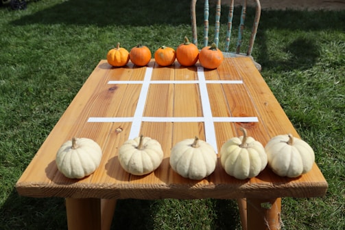 A wooden table outdoors with a tic-tac-toe grid made from white tape on the surface. There are small orange pumpkins at one end of the table and white pumpkins at the opposite end. The background consists of green grass and an old rustic chair is visible on one side of the table.