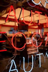 Neon lights display bright red and white letters against a darkened backdrop, likely the window of a store. Behind the glass, the soft glow illuminates a shop interior with shelves and possibly cosmetics or similar products visible.