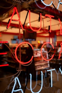 Neon lights display bright red and white letters against a darkened backdrop, likely the window of a store. Behind the glass, the soft glow illuminates a shop interior with shelves and possibly cosmetics or similar products visible.