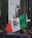 A person holds a large Mexican flag amidst a bustling city street lined with tall buildings. The surroundings include diverse buildings and a partially visible car hood, with people visible in the background.