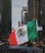 A person holds a large Mexican flag amidst a bustling city street lined with tall buildings. The surroundings include diverse buildings and a partially visible car hood, with people visible in the background.
