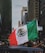 A person holds a large Mexican flag amidst a bustling city street lined with tall buildings. The surroundings include diverse buildings and a partially visible car hood, with people visible in the background.