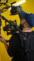 A person wearing a blue cap is operating a professional camera rig with a stabilizing setup. The background is bright yellow, and several camera accessories, including a monitor and a Ronin stabilizer, are visible.