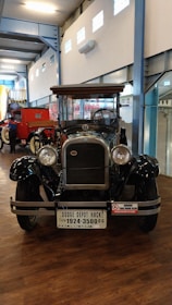 An antique car displayed indoors, with a polished black exterior and wooden accents. The vehicle has large, round headlights and a prominent grille. A license plate with 'Dodge Depot Hack' is attached to the front. The space has high ceilings and is part of a museum or exhibition hall, with bright lighting and other vintage vehicles nearby.