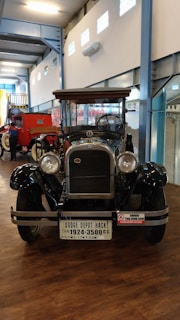 An antique car displayed indoors, with a polished black exterior and wooden accents. The vehicle has large, round headlights and a prominent grille. A license plate with 'Dodge Depot Hack' is attached to the front. The space has high ceilings and is part of a museum or exhibition hall, with bright lighting and other vintage vehicles nearby.