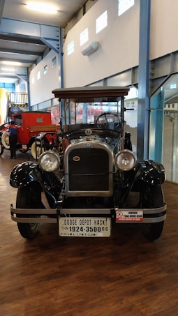 An antique car displayed indoors, with a polished black exterior and wooden accents. The vehicle has large, round headlights and a prominent grille. A license plate with 'Dodge Depot Hack' is attached to the front. The space has high ceilings and is part of a museum or exhibition hall, with bright lighting and other vintage vehicles nearby.
