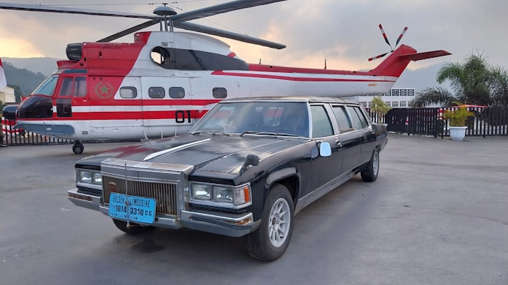A black limousine is parked on a paved area next to a red and white helicopter. The background shows a cloudy sky and some greenery, with a potted plant visible nearby.