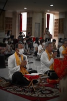 A group of people is seated in a room that resembles a meditation hall, with two prominent figures in white robes and orange scarfs sitting cross-legged on a mat. They appear to be in a meditative or prayerful posture, with one holding a microphone. Most individuals in the room are wearing face masks. The room is dimly lit with red curtains and decorative wall elements.