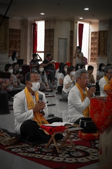A group of people is seated in a room that resembles a meditation hall, with two prominent figures in white robes and orange scarfs sitting cross-legged on a mat. They appear to be in a meditative or prayerful posture, with one holding a microphone. Most individuals in the room are wearing face masks. The room is dimly lit with red curtains and decorative wall elements.