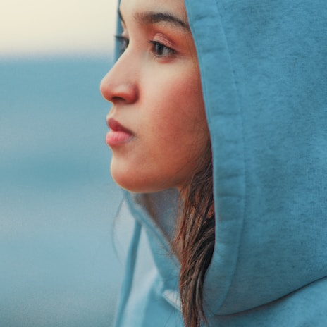 A person with a contemplative expression is seen in a close-up profile view. They are wearing a light blue hoodie, and their mood suggests introspection or daydreaming. The background is blurred and consists of soft blue hues.