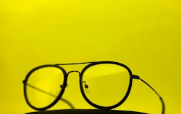 A pair of black-rimmed glasses with yellow-tinted lenses is placed against a bright yellow background. The glasses are centered in the image, drawing attention to their retro design and clean form.