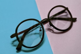 A pair of round black eyeglasses resting on a surface divided into two colors, light blue on the left and light purple on the right. The glasses cast a soft shadow, and the arms of the frames are folded.