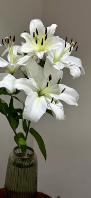 A bouquet of elegant white lilies placed in a ribbed green glass vase on a wooden surface. The lilies are in full bloom, showcasing large petals and prominent stamens. The background is a plain light wall, bringing focus to the flowers.