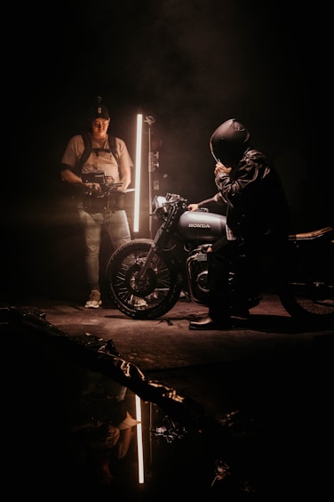 Two individuals are in a dimly lit setting with a vintage motorcycle. One person, equipped with filming gear, stands near a vertical light source, casting dramatic shadows. The other, seated on the motorcycle, appears to be adjusting a helmet.