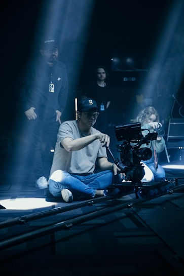 A group of people is engaged in a film or video production. One person sits on the floor operating a camera mounted on a dolly track, while another person, likely a director or assistant, stands nearby. The setting is dimly lit, with beams of light streaming down, creating a focused and dramatic atmosphere.
