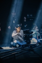 A group of people are on a film set with lighting creating dramatic shadows. A person wearing a cap and glasses is sitting on the floor operating a professional camera mounted on a dolly track. Other individuals are involved in setting up and possibly directing the scene, with one person holding a camera on the right side.