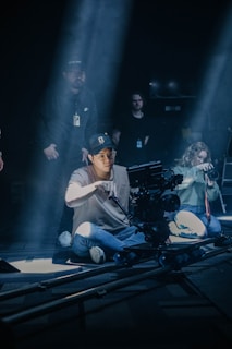 A group of people are on a film set with lighting creating dramatic shadows. A person wearing a cap and glasses is sitting on the floor operating a professional camera mounted on a dolly track. Other individuals are involved in setting up and possibly directing the scene, with one person holding a camera on the right side.