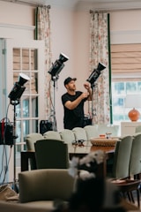 A person wearing a cap is adjusting lighting equipment in a room filled with furniture. The room features mint-green chairs, a wooden table, and floral curtains. Several professional lights are set up, suggesting a photography or video shoot setting. Natural light is coming in through a window with blinds, giving the room a bright and airy feel.