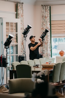 A person wearing a cap is adjusting lighting equipment in a room filled with furniture. The room features mint-green chairs, a wooden table, and floral curtains. Several professional lights are set up, suggesting a photography or video shoot setting. Natural light is coming in through a window with blinds, giving the room a bright and airy feel.