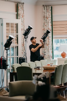 A person wearing a cap is adjusting lighting equipment in a room filled with furniture. The room features mint-green chairs, a wooden table, and floral curtains. Several professional lights are set up, suggesting a photography or video shoot setting. Natural light is coming in through a window with blinds, giving the room a bright and airy feel.