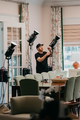 A person wearing a cap is adjusting lighting equipment in a room filled with furniture. The room features mint-green chairs, a wooden table, and floral curtains. Several professional lights are set up, suggesting a photography or video shoot setting. Natural light is coming in through a window with blinds, giving the room a bright and airy feel.