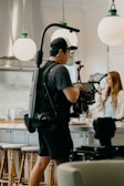 A camera operator wearing a cap and black outfit is filming a woman indoors. The man is using a professional camera setup, including a stabilizer and various filming equipment. The location appears to be a modern kitchen, with pendant lights, a marble backsplash, and bar stools visible. The woman, who has long hair, is seen in the background near the counter, possibly in conversation or preparing something.