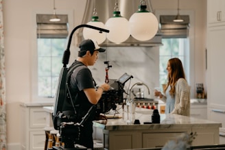 A person with camera equipment is filming in a well-lit modern kitchen. The kitchen features white cabinets, a marble island countertop, and three large spherical light fixtures hanging from the ceiling. A woman stands on the other side of the island, smiling and holding a cup.