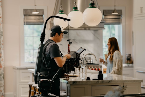 A person with camera equipment is filming in a well-lit modern kitchen. The kitchen features white cabinets, a marble island countertop, and three large spherical light fixtures hanging from the ceiling. A woman stands on the other side of the island, smiling and holding a cup.