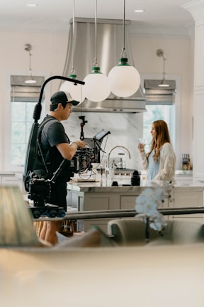 A person operates a professional camera on a rig in a bright, modern kitchen. Opposite them, a woman stands holding a glass. The scene is well-lit with large globe pendant lights overhead and stainless steel appliances visible in the background.