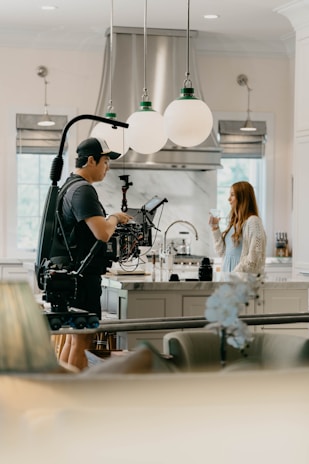 A person operates a professional camera on a rig in a bright, modern kitchen. Opposite them, a woman stands holding a glass. The scene is well-lit with large globe pendant lights overhead and stainless steel appliances visible in the background.