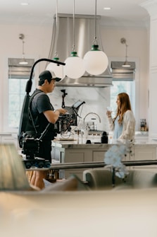 A person operates a professional camera on a rig in a bright, modern kitchen. Opposite them, a woman stands holding a glass. The scene is well-lit with large globe pendant lights overhead and stainless steel appliances visible in the background.