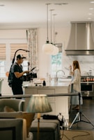 A film crew is in a kitchen setting, capturing a scene. A person operates a camera with stabilizing equipment while another person stands by the kitchen counter, drinking from a cup. The kitchen has modern decor with hanging lights, a stainless steel oven, and a marble countertop. Curtains cover the windows in the background.