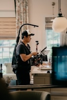 A person wearing a cap and a harness operates a professional camera rig inside a well-lit room. The room has modern decor with patterned curtains and a large pendant light hanging from the ceiling. The person is focused on the camera, which has multiple attachments and cables.
